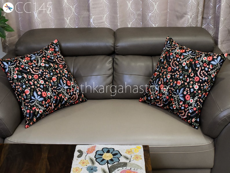 May include: Two black throw pillows with floral embroidery in shades of pink, blue, and white on a brown leather sofa. The pillows are arranged on the sofa with the floral patterns facing outward.