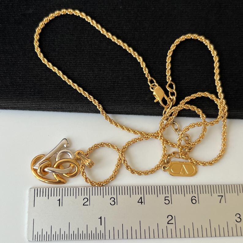 May include: A gold-toned necklace with a pendant featuring intertwined shapes in gold and silver. The chain is a twisted rope design. A ruler is placed next to the necklace, showing measurements in inches.
