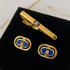 Puede incluir: Juego de gemelos y pinza de corbata de oro con esmalte azul y un monograma dorado. El monograma es un "JL" estilizado.
