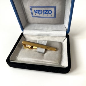 May include: A gold tie clip with the word "Kenzo" engraved on it, in a black velvet box with a blue lining and the word "Kenzo Paris" on the lid.