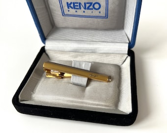 Könnte beinhalten: Ein goldener Krawattenclip mit der Gravur "Kenzo" in einer schwarzen Samtbox mit blauer Auskleidung und dem Schriftzug "Kenzo Paris" auf dem Deckel.