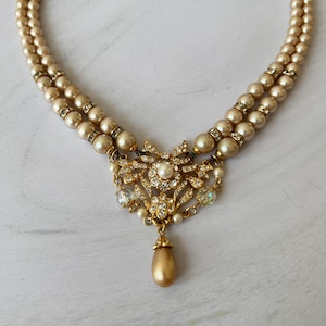 Puede incluir: Un collar de perlas de doble cadena con un colgante dorado con una perla y diamantes de imitación. El colgante tiene forma de flor.