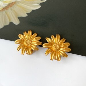 May include: A pair of gold-colored daisy earrings. Each earring features a detailed flower design with multiple petals and a textured center. The earrings are set against a white and black background.