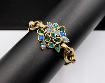 Puede incluir: Pulsera dorada con diseño floral. La pulsera presenta un grupo de piedras preciosas azules y verdes de varias formas. Los eslabones de la cadena son gruesos y texturizados. La pulsera se exhibe sobre un objeto cilíndrico negro.