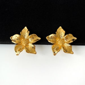 May include: A pair of gold-tone flower-shaped earrings. The earrings are made of metal and have a textured surface.