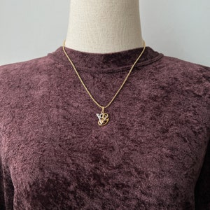 May include: Gold-toned necklace with a pendant featuring the letters "VS" intertwined with a heart design. The pendant has a gold outline with a silver-colored interior. The necklace is displayed on a dark burgundy velvet top.