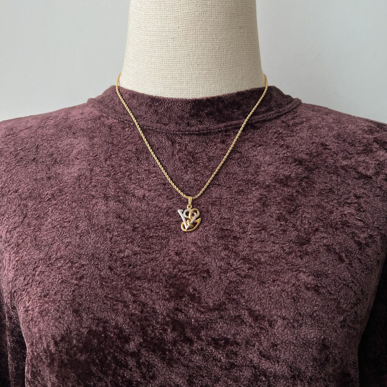 May include: Gold-toned necklace with a pendant featuring the letters "VS" intertwined with a heart design. The pendant has a gold outline with a silver-colored interior. The necklace is displayed on a dark burgundy velvet top.