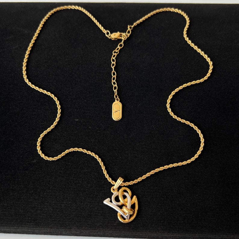 May include: Gold-toned necklace with a rope chain and a pendant featuring the letters "V" and "L" in a stylized design. The pendant has gold and silver elements. The necklace is displayed on a black surface.