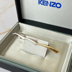 May include: A silver and gold tie clip with the word "KENZO" engraved on the gold portion.