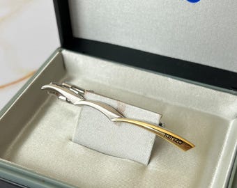 Könnte beinhalten: Ein silberner und goldener Krawattenclip mit dem Wort "KENZO" auf dem goldenen Teil.