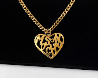 Puede incluir: Un collar de cadena dorada con un colgante en forma de corazón. El colgante está recortado con las letras "MOSCHINO" en su interior.