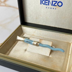 May include: A light blue tie bar with the word "KENZO" in silver lettering. The tie bar is in a black box with a white interior and the word "KENZO HOMME" printed on the top.