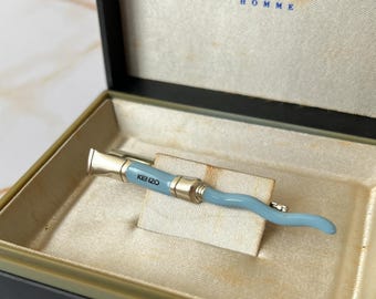 Könnte beinhalten: Ein hellblauer Krawattenclip mit dem Wort "KENZO" in silbernen Buchstaben. Der Krawattenclip befindet sich in einer schwarzen Box mit weißem Innenraum und dem aufgedruckten Wort "KENZO HOMME" auf der Oberseite.