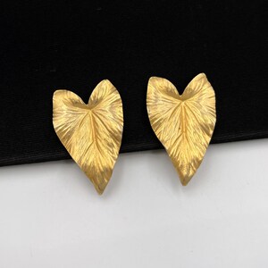 May include: A pair of gold-colored leaf-shaped earrings. The earrings have a textured surface with detailed veining, resembling real leaves. The earrings are set against a black and white background.
