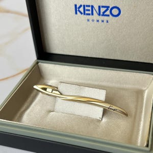 May include: Gold tie bar with a curved design and the word "Kenzo" engraved on it. The tie bar is in a black box with a white interior and the word "Kenzo Homme" printed on the top.