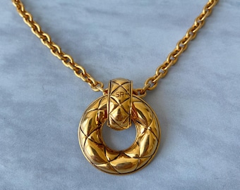 Puede incluir: Un collar de cadena de oro con un gran colgante redondo. El colgante tiene un diseño acolchado y una pequeña inscripción "SR".