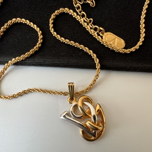 May include: Gold-toned necklace with a pendant featuring a stylized design in gold and silver. The chain is a twisted rope style, and the pendant has a gold bail. The necklace is displayed against a black background.