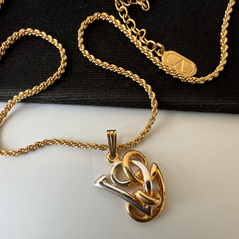 May include: Gold-toned necklace with a pendant featuring a stylized design in gold and silver. The chain is a twisted rope style, and the pendant has a gold bail. The necklace is displayed against a black background.