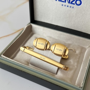 May include: A gold-colored cufflink set with a tie clip. The cufflinks and tie clip are engraved with the word "KENZO". The set is displayed in a black box with a white interior.
