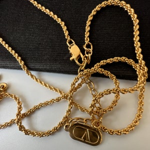 May include: A gold-colored necklace with a rope chain design. The necklace features a lobster clasp closure and a small, rectangular pendant with a stylized "V" logo. The jewelry is displayed against a black background.