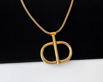Puede incluir: Un collar dorado con un colgante grande. El colgante presenta una barra vertical que cruza una forma redondeada, creando un diseño estilizado. La cadena es de estilo tejido, sobre un fondo en blanco y negro.