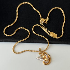 May include: Gold-toned necklace with a rope chain and a pendant featuring a gold and silver design. The pendant has a stylized shape with intertwined elements. The necklace is displayed on a white and black surface.