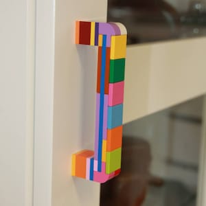 Könnte beinhalten: Ein farbenfroher Schrankgriff mit geometrischem Design. Der Griff besteht aus gestapelten rechteckigen Blöcken in verschiedenen Farben, darunter Rot, Orange, Gelb, Grün, Rosa und Blau. Der Griff ist an einer weißen Schranktür befestigt.