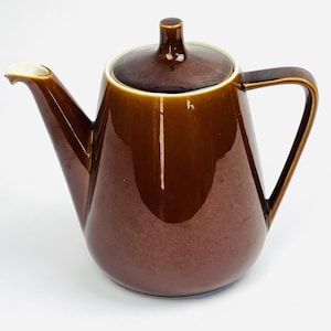 Op de afbeelding: Een glanzend bruine keramische theepot met bijpassend deksel, voorzien van een taps toelopend lichaam, een gebogen handvat en een tuit met een witte binnenrand. De theepot heeft een gladde, reflecterende afwerking en een functioneel ontwerp.
