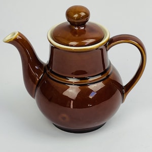 Peut inclure: Une théière en céramique marron foncé brillant avec un corps arrondi, une anse incurvée et un couvercle assorti doté d'un bouton. Le bord du couvercle et l'extrémité du bec verseur sont soulignés par un fin liseré crème contrastant.