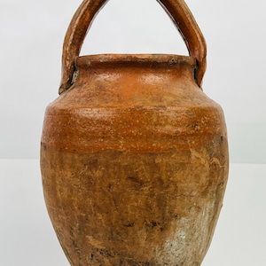 Peut inclure: Une cruche en poterie de style antique avec une anse. La cruche présente un corps arrondi et une large ouverture, avec une anse incurvée. La couleur est un mélange de teintes marron et orange, avec des signes d'usure et de vieillissement.
