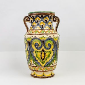 Op de afbeelding: Een decoratieve keramische vaas met een crèmekleurige basis en ingewikkelde handgeschilderde ontwerpen. De vaas heeft een levendig kleurenpalet van geel, blauw, groen en bruin, met twee gebogen handvatten. De vaas is ongeveer 25 cm hoog.