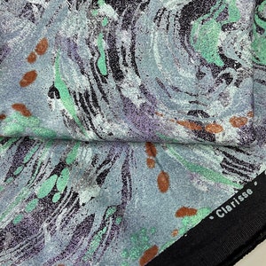 May include: A close-up of a fabric with a swirling, marbled pattern in shades of gray, green, and black. The fabric has a textured surface and small brown spots. The fabric has a black border with the text "Clarisse &copy; MCMLXXXVIII EDITE PAR Taco" printed on it.