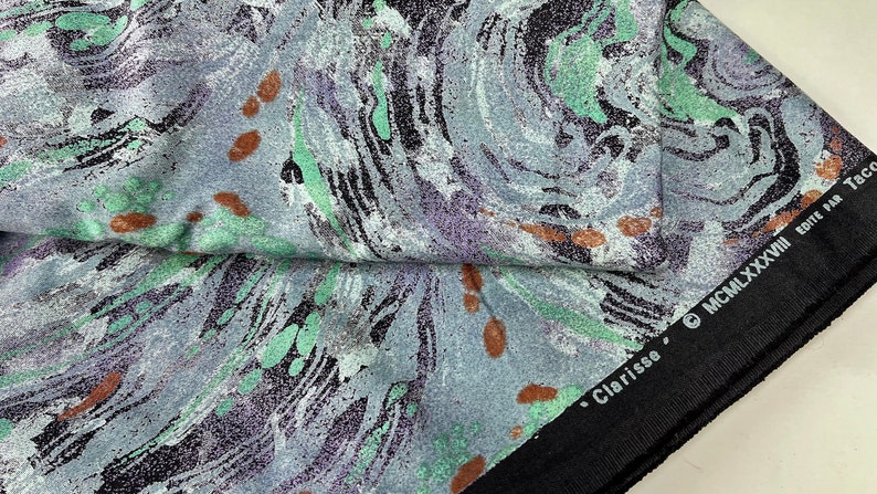 May include: A close-up of a fabric with a swirling, marbled pattern in shades of gray, green, and black. The fabric has a textured surface and small brown spots. The fabric has a black border with the text "Clarisse &copy; MCMLXXXVIII EDITE PAR Taco" printed on it.