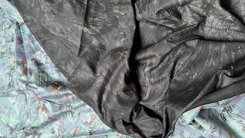 May include: A close-up of two fabric swatches. One is a black fabric with a subtle marbled pattern. The other is a light blue fabric with a marbled pattern in shades of green, purple, and orange.
