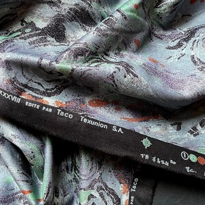 May include: Close-up of a blue and green fabric with a marbled pattern. The fabric has a black border with the text "Clarisse" and "Edite par Taco Texunion S.A."