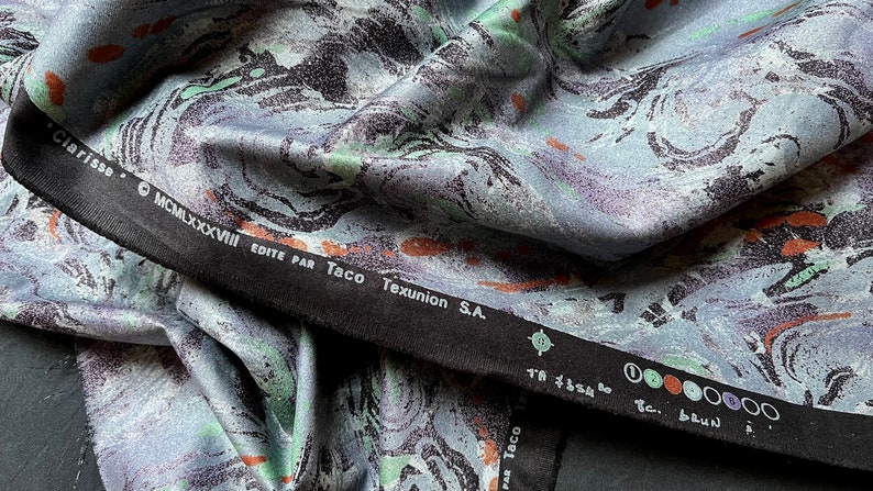 May include: Close-up of a blue and green fabric with a marbled pattern. The fabric has a black border with the text "Clarisse" and "Edite par Taco Texunion S.A."
