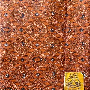 以下が含まれることがあります： 白と黒の幾何学模様が繰り返し描かれた茶色の布。布には、黄色の背景に白い文字で「Batik Cap Nori」と「Rotong Panjang」と書かれたラベルが付いています。