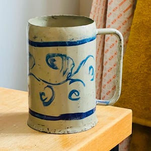 Op de afbeelding: Een bierpul in vintage-stijl met een crèmekleurige body en blauwe decoratieve krullen en banden. De mok heeft een grote handgreep en staat op een houten oppervlak.
