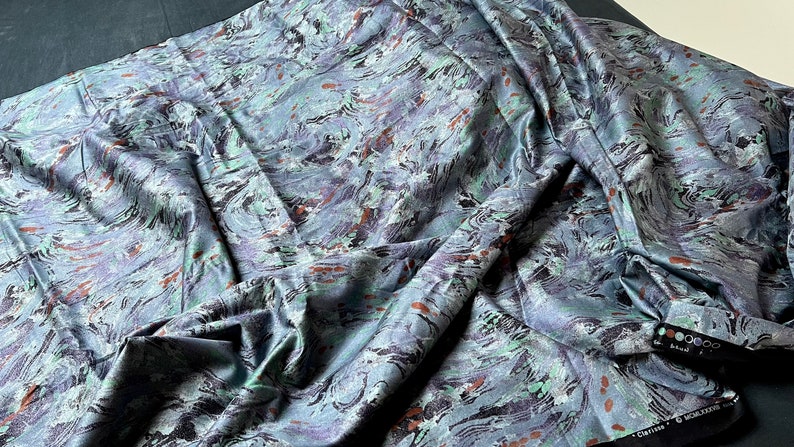 May include: A close-up of a blue, green, and brown fabric with a marbled pattern. The fabric is folded and draped, showing the texture and detail of the design.
