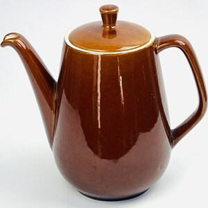 Könnte beinhalten: Eine glänzende, dunkelbraune Kaffeekanne aus Keramik mit passendem Deckel, einem geschwungenen Griff und einem langen, spitz zulaufenden Ausguss. Die Kanne hat eine glatte, reflektierende Oberfläche und ein schlichtes Design mit weißem Rand.