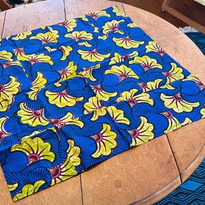 Op de afbeelding: Een vierkant stuk stof met een opvallend bloemmotief in blauw, geel en rood. De stof ligt op een houten tafel, met een schaar in de buurt. Het patroon toont gestileerde bloemen.