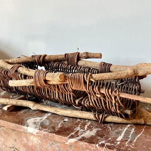 May include: A rustic woven basket made from natural branches and wire. The basket is brown and has a rectangular shape. It is sitting on a marble surface.