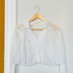 Op de afbeelding: Een witte blouse met lange mouwen, een V-hals en een knoopsluiting, hangend aan een houten hanger. De blouse heeft een licht doorschijnende uitstraling en is gemaakt van een lichte stof. De achtergrond is een witte muur met een houten deurkozijn.