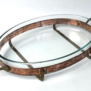 Peut inclure: Un plat de cuisson ovale en verre inséré dans un cadre en cuivre patiné. Le support présente deux poignées latérales incurvées, une bande métallique texturée sur le pourtour et quatre petits pieds qui surélèvent le récipient en verre transparent.