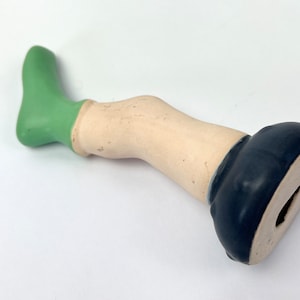 Może przedstawiać: Ceramiczna figurka nogi z zieloną skarpetą i czarnym butem. Noga jest zgięta w kolanie, a but znajduje się u dołu figurki.