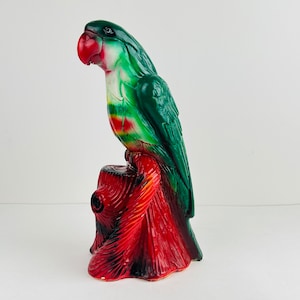 May include: A green and red parrot figurine perched on a red and black tree stump. The parrot has a red beak and white markings on its chest.