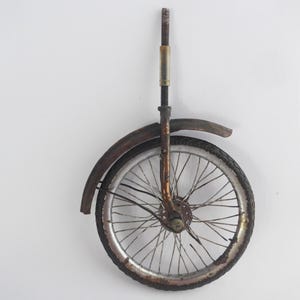 May include: A rusty bicycle wheel with a metal spoke and a brown tyre. The wheel is mounted on a white wall.
