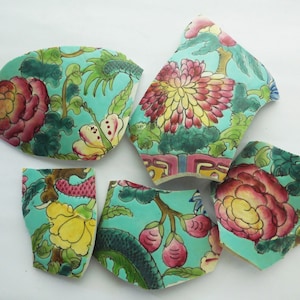 May include: Six pieces of broken turquoise blue ceramic with floral patterns in shades of pink, yellow, and green. The pieces are arranged on a white surface.