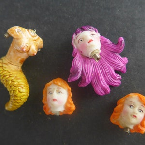 Op de afbeelding: Vijf kleine plastic figuurtjes van vrouwenhoofden met verschillende haarkleuren en -stijlen. De figuurtjes dragen allemaal witte jurken en hebben geschilderde gezichten.