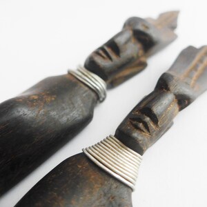 May include: Two dark brown, carved wooden figures with human faces and silver wire accents around the neck. The figures have a weathered appearance and are set against a white background.